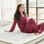 A woman in maroon pajamas sits smiling on an Avocado Green Ultra Plush Mattress by Avocado Green Mattress in a bright room with large windows.