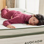 A woman wearing pink pajamas lies comfortably on her stomach atop a white Avocado Green Ultra Plush Mattress with green trim. She appears relaxed and is smiling with her eyes closed, enjoying the luxurious comfort of the Avocado Green Mattress brand.
