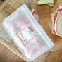 A (re)zip 2 Gallon + 2 Half Gallon reusable bag filled with sliced deli meat is on a wooden table beside a sandwich—ideal for meal prep or storing leftovers.