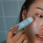A person applies the Baeo Bare Face - Soothing Face Stick with a palm tree logo to their cheek, hydrating sensitive skin, as part of their face appears against a tiled background.