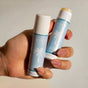 A hand with manicured nails holds two Baeo Bare Face - Soothing Face Sticks with light blue labels and a white palm tree; one stick is closed and the other shows the hydrating product—ideal for sensitive skin.