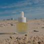A small glass dropper bottle labeled Baeo Bare Glow – Radiant Face Oil stands upright on sandy beach under a blue sky with scattered clouds, promising restorative hydration and a glowing complexion.