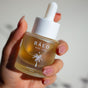 A hand with manicured nails holds a Baeo Bare Glow - Radiant Face Oil bottle, featuring a white cap and palm tree design—a lightweight face oil perfect for glowing skin.