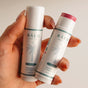 A hand holds two Baeo Bare Tint - Mineral Lip + Cheek Tint tubes—one open, revealing pink hydrating blush, and one closed. Both feature a white label with a palm tree illustration.