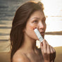 At sunset by the beach, a woman applies Baeo’s Bare Tint - Mineral Lip + Cheek Tint in peach to her cheeks, as gentle sunlight softly lights up her face.