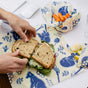 Someone wraps a sandwich in Bees Wrap Lunch Pack at a desk, while another eco-friendly wrap holds baby carrots. Blue animal and plant prints decorate the wraps. Apple slices, a glass of water, and a keyboard are also on the desk.