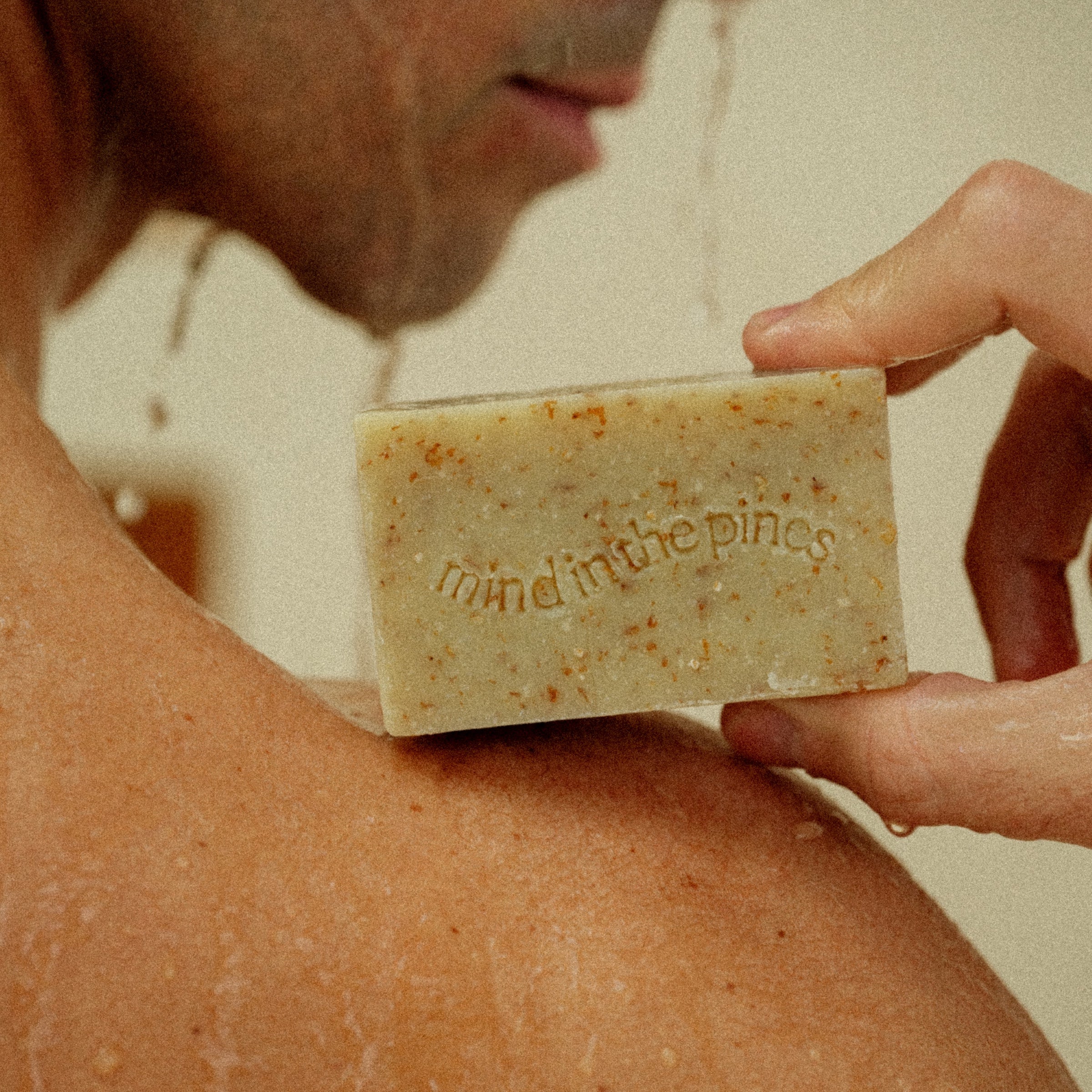 A person in the shower holds Jackfir’s The Bushwhacker Bar Soap against their wet shoulder, its “mind in the pines” engraving and water droplets highlighting the soap’s all-natural ingredients.