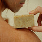 A person in the shower holds Jackfir’s The Bushwhacker Bar Soap against their wet shoulder, its “mind in the pines” engraving and water droplets highlighting the soap’s all-natural ingredients.