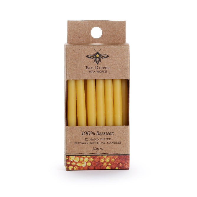 A brown box labeled Big Dipper Wax Works contains 12 yellow, all-natural Birthday Tapers. The packaging has a bee logo, a honeycomb pattern at the bottom, and a clear window displaying the candles.