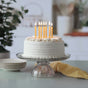 A white frosted cake shines with eight lit Big Dipper Wax Works Birthday Tapers (12 pk) atop a glass stand, while stacked plates and a yellow striped cloth set the scene for an all-natural celebration in a cozy kitchen.