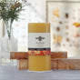 A large 100% Pure Beeswax Pillar Candle by Big Dipper Wax Works, featuring a bee label, stands on a table with books and a vase of yellow flowers, adding eco-friendly ambiance to a toxin-free home. Botanical prints are softly blurred in the background.