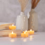Several Pure Beeswax Tea Lights Natural by Big Dipper Wax Works are displayed on a white surface and books, with ceramic vases and dried grass behind them. The softly lit scene feels cozy and calm, enhanced by their gentle natural aroma.