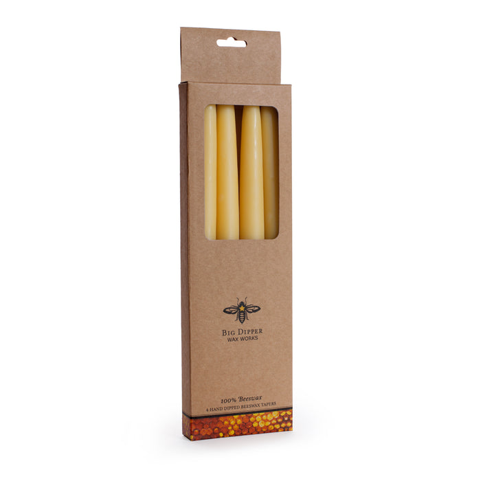 A brown box with a clear window showcases four yellow Hand Dipped Tapers by Big Dipper Wax Works. The box features a bee illustration, the brand name, and “100% Beeswax,” highlighting these natural, toxin-free candles.