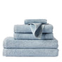 A stack of neatly folded light blue Coyuchi Air Weight® Organic Towels is arranged against a plain white background. One small organic cotton towel from the collection is draped over the edge of the stack.