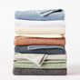 A stack of seven neatly folded Coyuchi Air Weight® Organic Towels in a variety of colors—blue, white, beige, rust, green, and gray—arranged on a white background. Expertly crafted from absorbent organic bath towel fabric for exceptional softness and functionality.