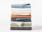 A stack of eight neatly folded Coyuchi Air Weight® Organic Towels in an assortment of colors—blue, white, beige, rust, gray, green, and dark gray—arranged on a white background.