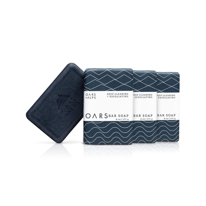 Four dark blue rectangular bars of Oars + Alps Exfoliating Bar Soap are shown, three in patterned wrappers and one unwrapped displaying the brand logo.