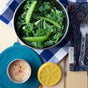 A metal bowl with fresh kale and snap peas is beside utensils on a blue plaid napkin. Nearby, an EcoLunchBox Seal Cup Mini with creamy pepper dressing and a yellow lid sits on the wooden surface.