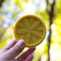 A hand holds a round, yellow object like a lemon slice against a blurred outdoor background, similar to showcasing the EcoLunchBox Seal Cup Mini container outdoors.