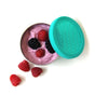 The EcoLunchBox Seal Cup Solo, a round stainless steel container with a turquoise lid, is filled with pink yogurt topped with raspberries and blackberries. Three raspberries are placed beside it on a white background.