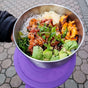 An EcoLunchBox Seal Cup Jumbo stainless steel container filled with poke ingredients—diced fish, avocado, seaweed salad, radish, kimchi, green onions, and sesame seeds—held above a purple surface with a patterned stone walkway in the background.