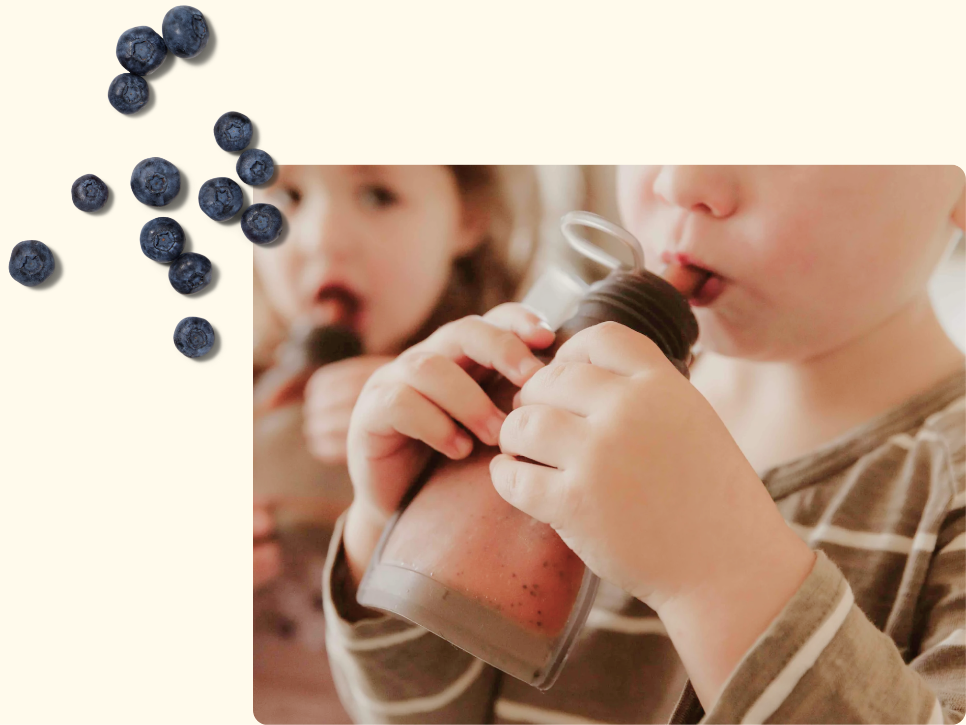 Two young children drink smoothies, one holding a bottle with both hands. Blurred in the background, the other child sips from a cup. Blueberries are scattered on the upper left of the light background.