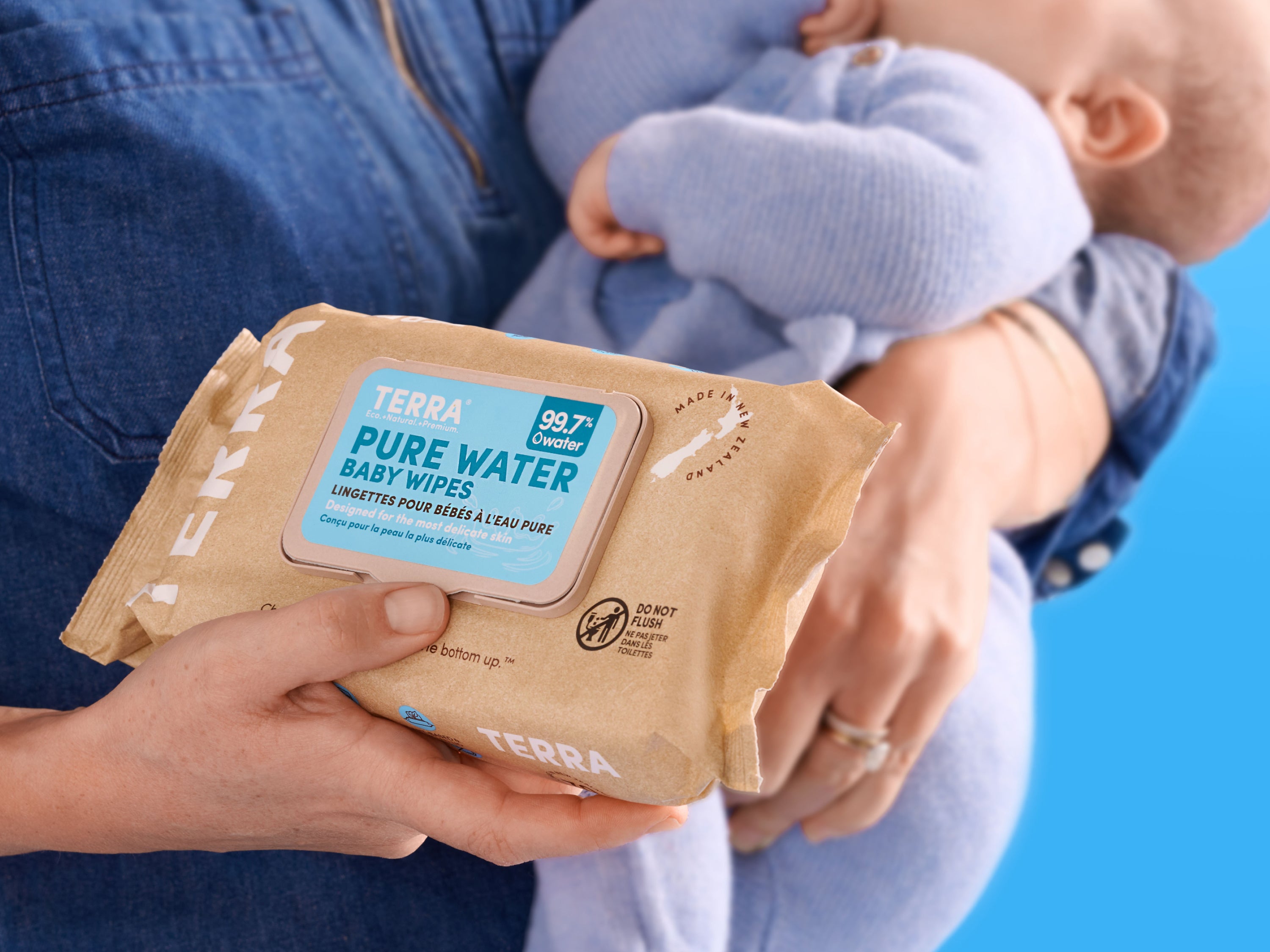 A person holds a package of TERRA Pure Water baby wipes while cradling a baby in their other arm, both wearing blue clothing, against a blue background.