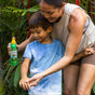 A woman smiles as she sprays Badger Balm’s Anti-Bug Shake & Spray - Mosquito Repellent onto a young boy’s arm in a lush garden. The boy, dressed in a blue shirt, looks down at his arm while the DEET-free bug spray is applied amidst green plants.