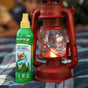 A green bottle of Badger Balm Anti-Bug Shake & Spray - Mosquito Repellent stands next to a lit red lantern on a plaid surface, with a blurred background.