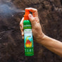 A hand sprays a green bottle labeled Badger Balm Anti-Tick Repellent Shake & Spray, an organic, DEET-free tick repellent, against a brown outdoor background.