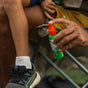 An adult sprays Badger Balm Anti-Tick Repellent Shake & Spray onto a child’s leg. The child, sitting on a folding chair, is wearing a white sock and a black and blue sneaker.