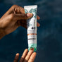 A hand dispenses a small amount of Badger Balm Tinted Face Mineral Sunscreen, SPF 40 - Sheer Tint onto a fingertip, set against a blurred dark blue background.