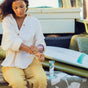A woman sitting on a patterned blanket applies lotion to her arm next to a surfboard and a tube of Badger Balm Daily Mineral Sunscreen - SPF 50, with a vehicle in the background.