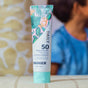 A tube of Badger Balm Daily Mineral Sunscreen - SPF 50, a reef-safe mineral sunscreen, featuring a tropical leaf design, stands on a striped surface with a blurred person in a blue shirt visible in the background.