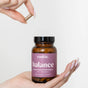 A hand holds a brown Rookie Wellness Balance Capsules bottle, while another holds a capsule above it. The label notes hormone balance and PMS support, featuring dill and chasteberry for monthly wellness.