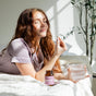 A woman with long red hair sits on a sunlit bed, eyes closed and smiling, holding a glass of water and a Balance Capsules - Hormone Balance + PMS Support capsule by Rookie Wellness. The bottle promises comfort and tranquility beside her.