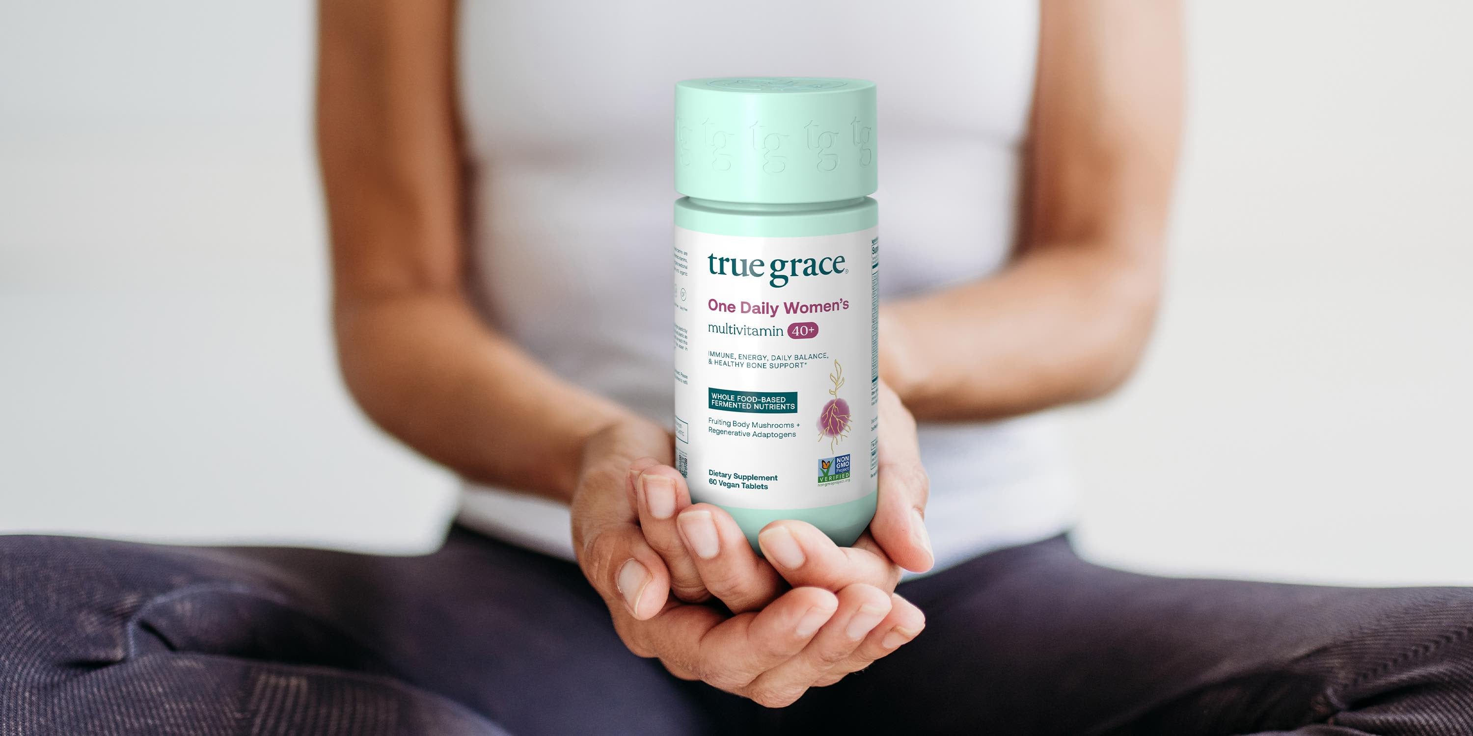 A person seated cross-legged holds a bottle of True Grace One Daily Women’s Multivitamin Ultra in their hands, with the label clearly visible.