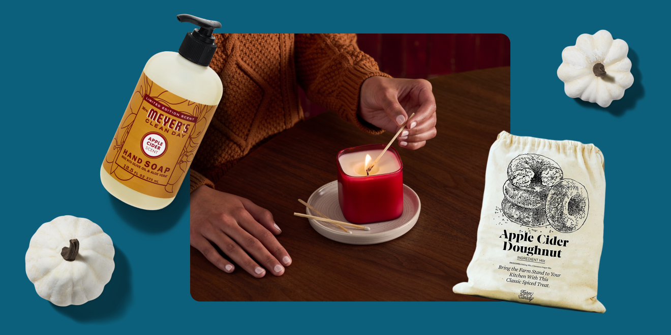 Hand holding a lit candle with a bottle of soap, a bag of apple cider doughnut, and white pumpkins on a blue background.
