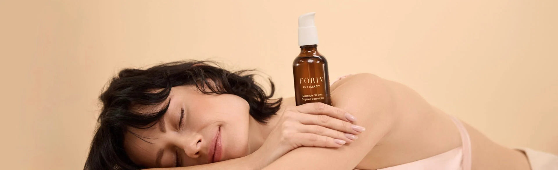 Woman holding a bottle of Foria Massage Oil on a beige background