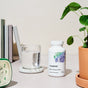 A bottle of Thorne Basic Nutrients 2/Day rests on a light table next to a glass of water, green clock, stack of books, and potted plant against a neutral backdrop.