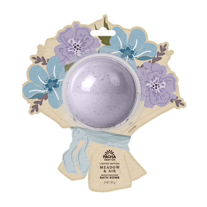 A round lavender bath bomb is packaged in a bouquet-shaped cardboard with illustrated purple and blue flowers and a blue ribbon. The label says Pacha Soap Co. Bath Bomb Card - Meadow & Air.