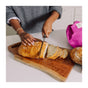 A person in a gray sweater slices a loaf of bread on a wooden cutting board, with several slices already cut. A Bee's Wrap Artisan Bread Bag is visible nearby on the counter for reusable storage.