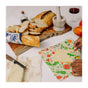 A table with bread, a loaf, cheese, tomatoes, red wine, and hands wrapping cheese in Bee's Wrap Beeswax Wraps for Food—Cut to Size Roll: a sustainable sandwich wrap and perfect plastic wrap alternative.