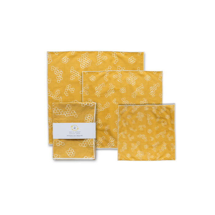 Four yellow reusable gift wraps with a white honeycomb pattern—three unfolded, one folded with a label—are displayed against a white background, featuring Bee's Wrap Reusable Gift Wrap Set made from certified organic cotton.