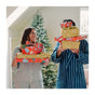 A woman and a man in pajamas stand by a Christmas tree, smiling at each other while holding gifts wrapped in Bee's Wrap Reusable Gift Wrap Set - Certified Organic Cotton. The man’s face is partly hidden behind the presents.