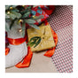 A gift wrapped in Bee's Wrap Reusable Gift Wrap Set—Certified Organic Cotton—rests by a Christmas tree, near red ribbon, other presents on a white knit stocking, and a checkered rug.