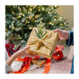 Two people exchange a gift wrapped in Bee's Wrap Reusable Gift Wrap Set—certified organic cotton—adorned with a pine branch, in front of a decorated Christmas tree with presents underneath.