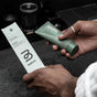 A person wearing silver rings and a white robe holds a green tube of before Classic Purifying Toothpaste w/ Fluoride, Anti-Cavity and its matching box above a dark bathroom countertop.