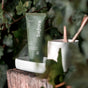 A green tube of before Classic Purifying Toothpaste w/ Fluoride, Anti-Cavity stands upright on a white tray outdoors among lush leaves. A white cup with two wooden sticks sits beside it, highlighting the natural, fresh ambiance.
