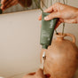 Someone squeezes before Classic Purifying Toothpaste w/ Fluoride, Anti-Cavity from a green tube onto a toothbrush, with a beige vase and mirror in the background.